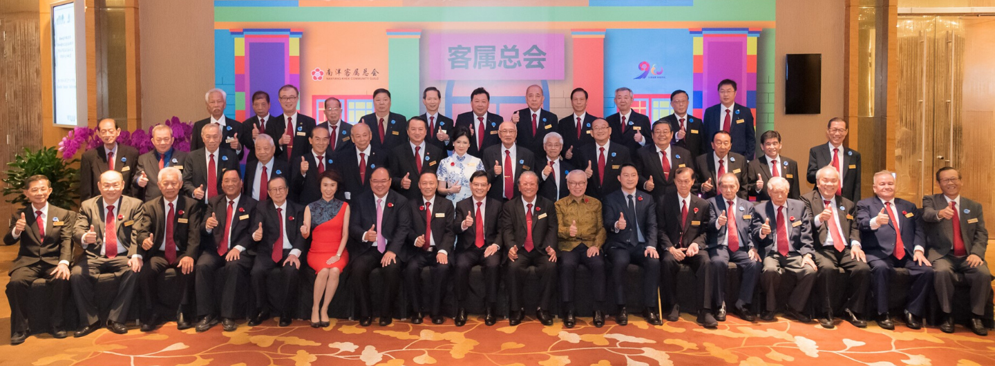 Group portrait of people in suits, some giving thumbs up, with a Nanyang Hakka Community Guild banner.